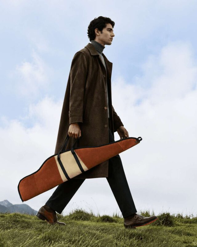 A stylish man in a brown coat carries a Frontier Leather Co. bag while walking through a scenic outdoor landscape.